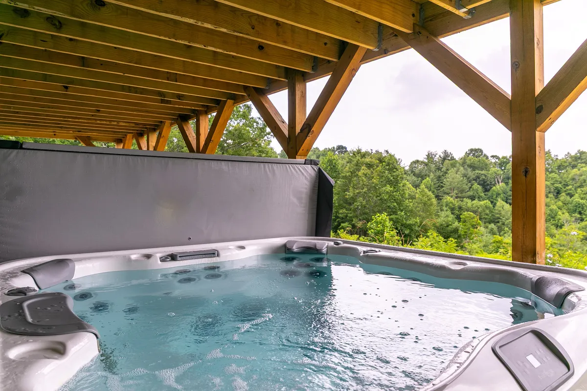 A serene hot tub set on a wooden deck, framed by sturdy beams and surrounded by lush green scenery. The scene is peaceful, hinting at sunset or starlit evenings with views that stretch out to the mountains.