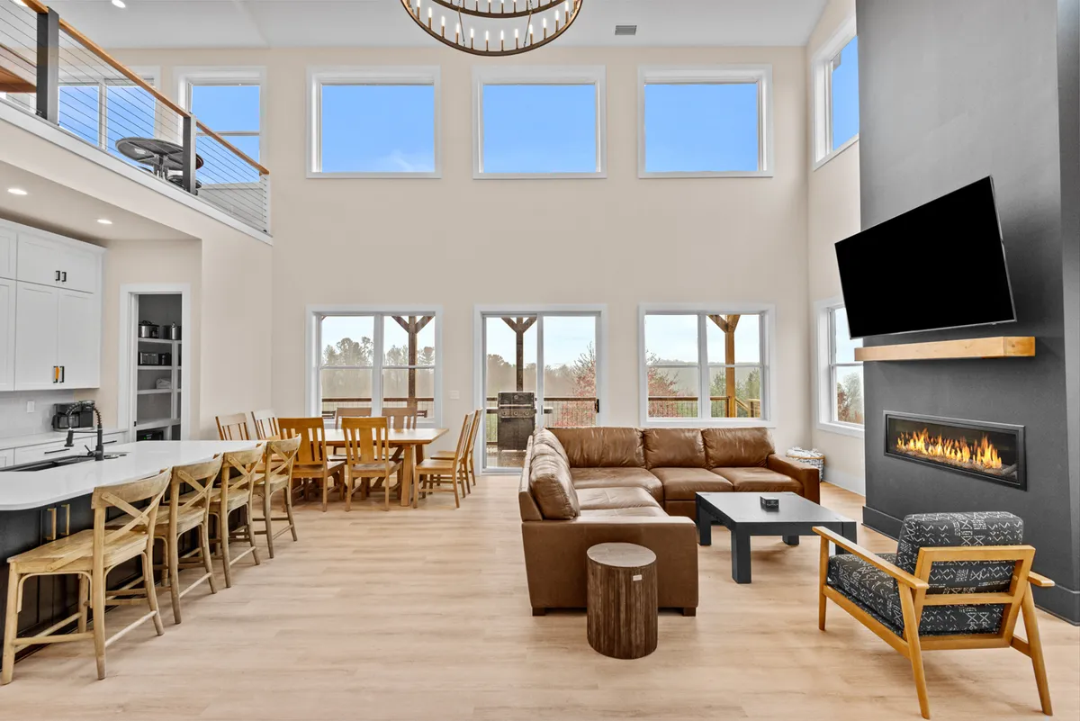 A spacious open-concept living area with a large sectional sofa facing a modern fireplace, with a dining area and chandelier visible. Expansive windows provide sweeping views and fill the space with natural light, while premium finishes and stylish decor create a warm, inviting atmosphere. The seamless flow between living and dining spaces encourages gatherings and relaxation.