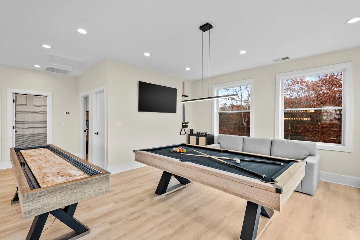This image captures a vibrant game room featuring a pool table, shuffleboard table, plush sofa, large windows showcasing mountain views, and a television. The scene radiates a playful and relaxed vibe with ample space for multiple activities, highlighting both the entertainment amenities and the inviting atmosphere.