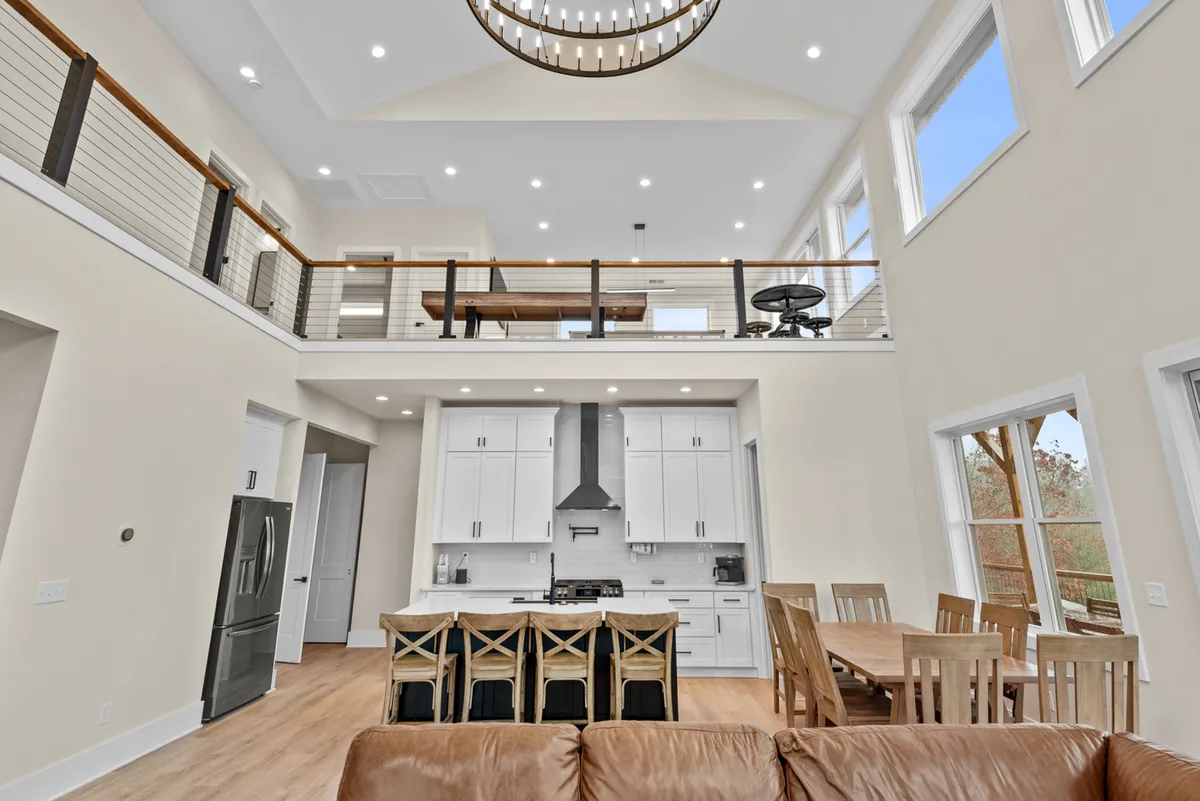A modern open kitchen and dining area featuring a large chandelier, high ceilings, a spacious dining table, and an expansive kitchen island. The area is bathed in warm light and showcases premium appliances and finishes, with ample seating for group meals and celebrations.