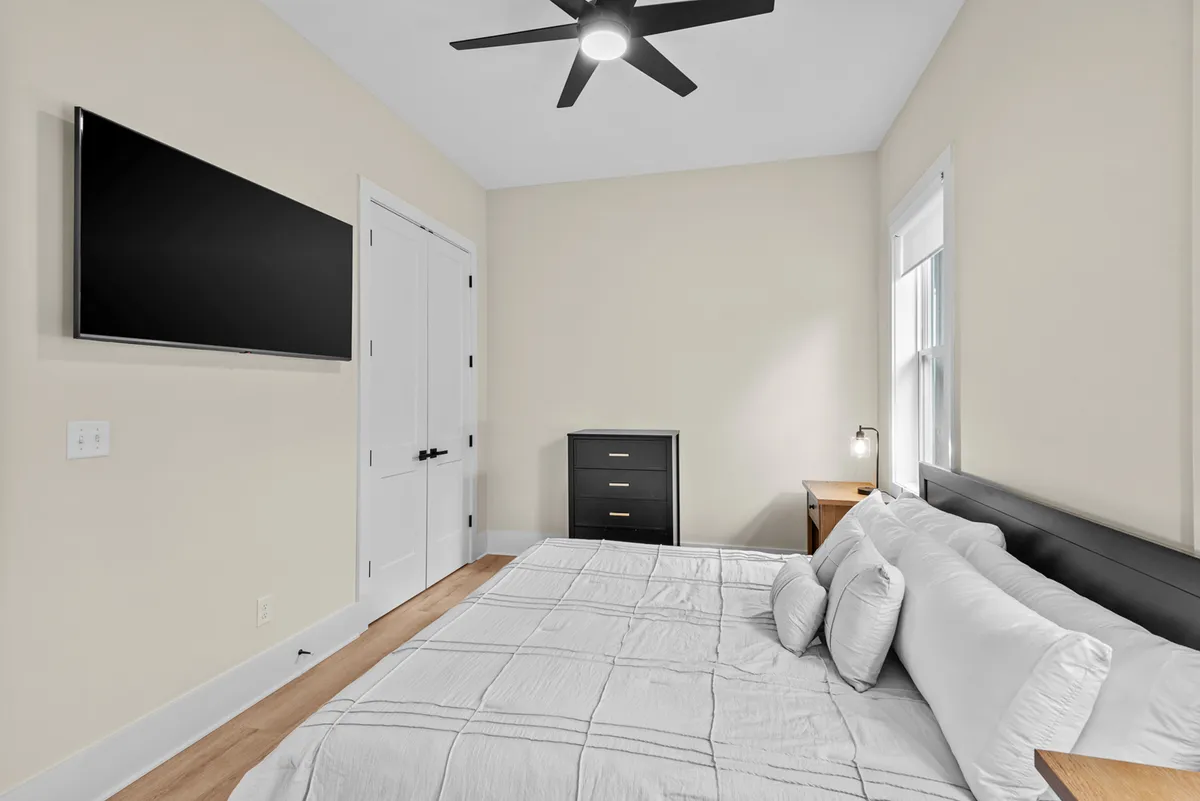A tranquil and inviting bedroom with a cozy bed, an elegant black dresser, a wall-mounted TV, and a sunlit window. The soft hues and uncluttered décor foster a relaxing environment ideal for unwinding.