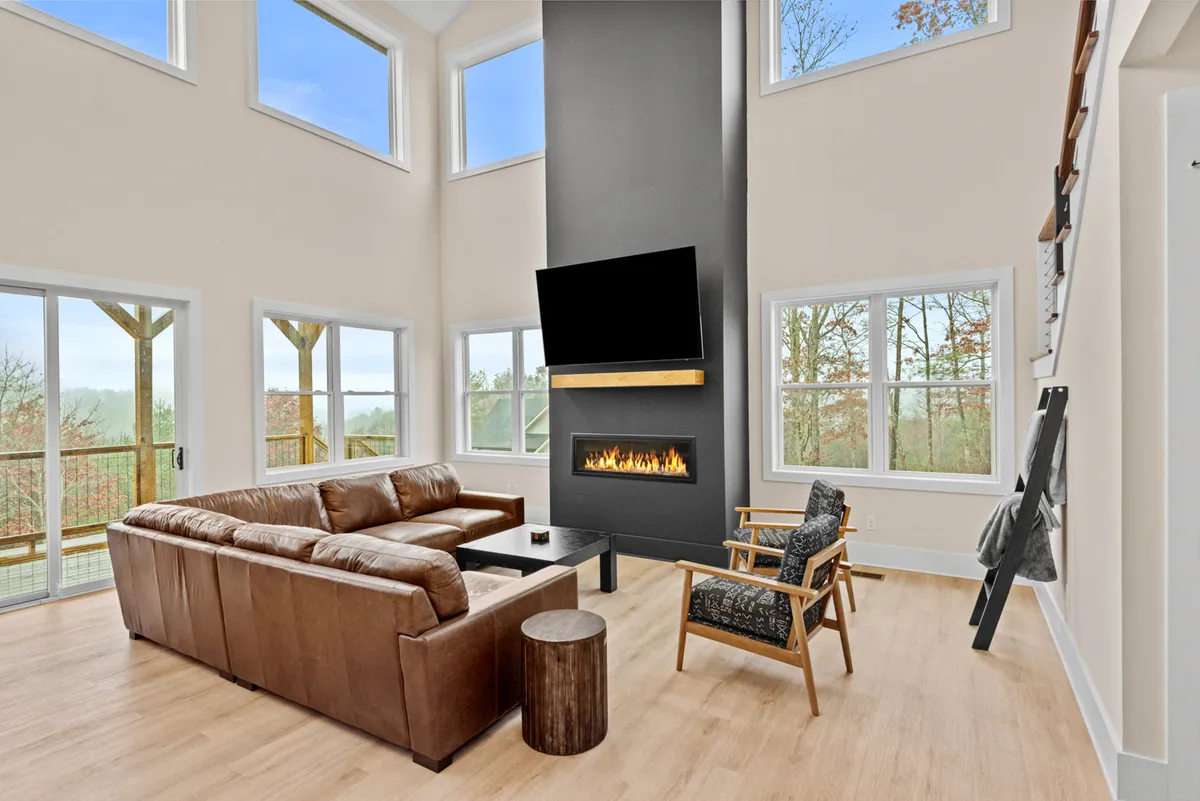 A contemporary living area with a large sectional sofa, modern fireplace, and floor-to-ceiling windows revealing scenic views. The space exudes warmth and comfort, ideal for relaxing or socializing as the setting sun fills the room with a golden glow.