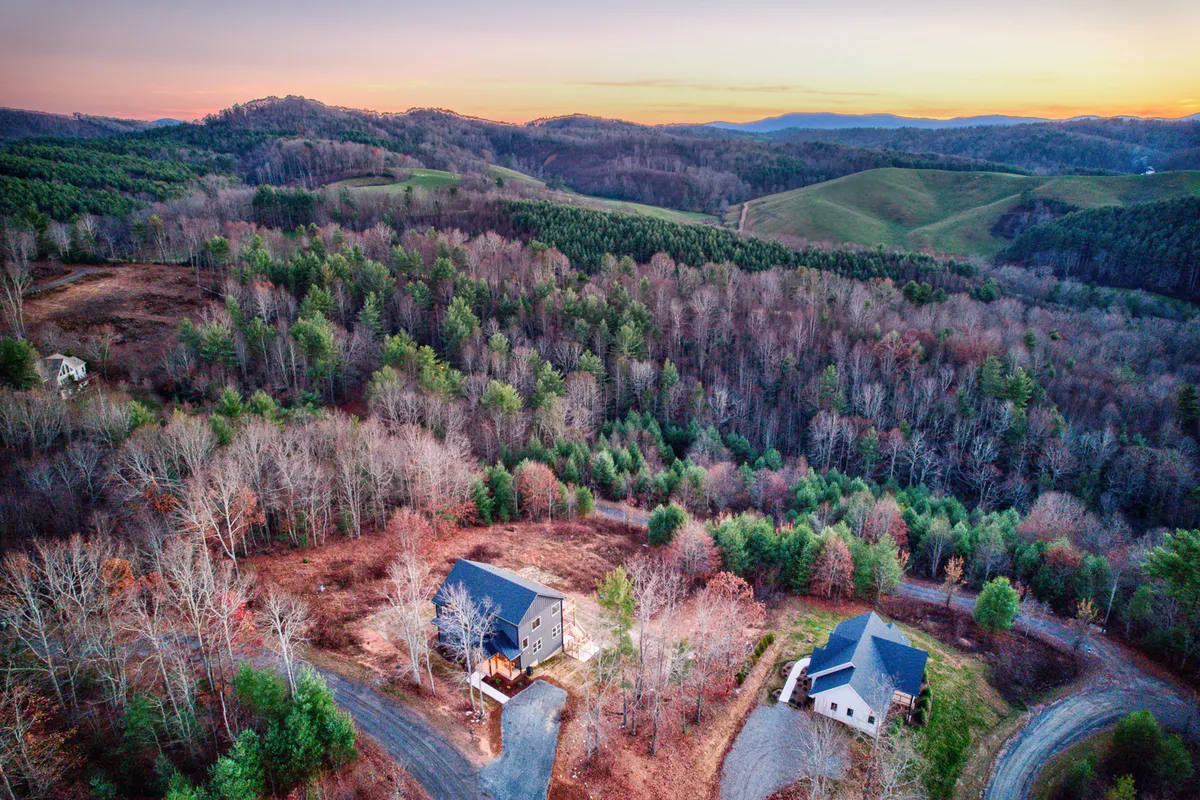 A wide-angle view of two houses nestled among rolling hills and dense forest, capturing an expansive landscape ideal for hiking and nature walks. The image exudes tranquility with sunset hues washing over the trees and open spaces, evoking the sense of calm after outdoor adventures.