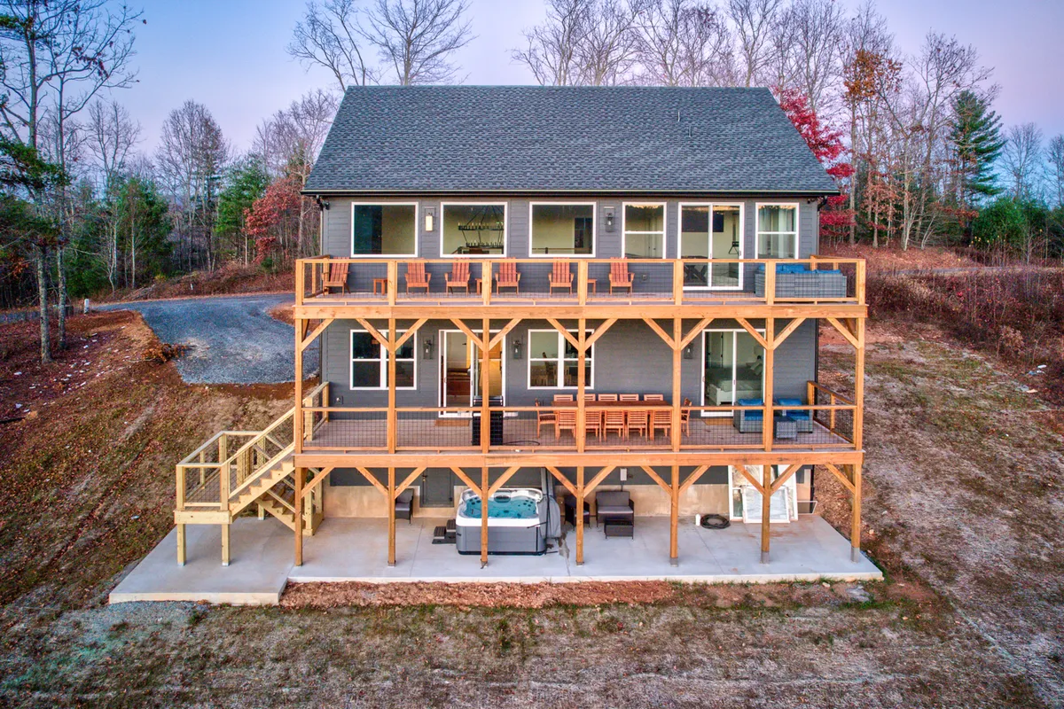 A sweeping view of multi-level decks featuring a comfortable seating area, a bubbling hot tub with mountain vistas, and large windows connecting to the interior. Guests are seen dining outdoors, relaxing in the hot tub, and gathering by the fire pit as the sun sets, all surrounded by a manicured yard and the Blue Ridge landscape.
