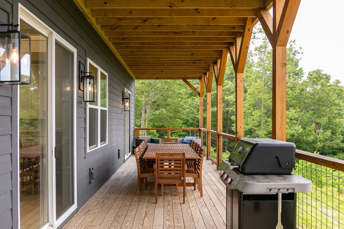 An outdoor dining area featuring a spacious table and grilling station, with a wooden railing overlooking lush greenery. The setup invites groups to enjoy al fresco meals and barbecue feasts in a scenic setting.