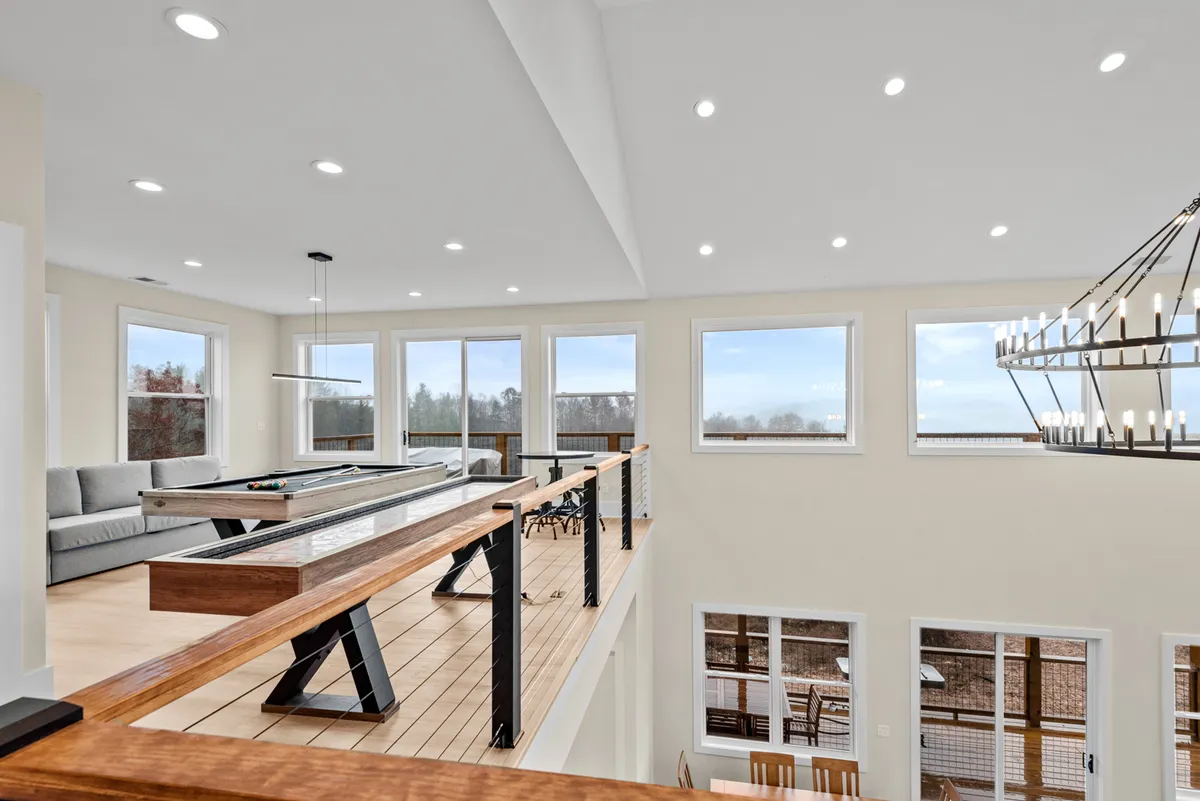 A stylish upper-level loft with a pool table, comfortable sofa, large windows flooding the room with natural light, and a balcony railing overlooking the main living space. The mountain scenery beyond the windows adds to the resort-like feeling.