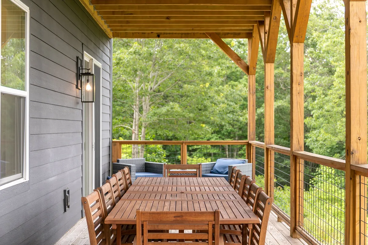 A large wooden dining table on the deck, surrounded by comfortable seating, with the lush greenery of the landscape in the background. The space is styled for group gatherings, highlighting sunset dinners, family celebrations, or quiet mornings with coffee.