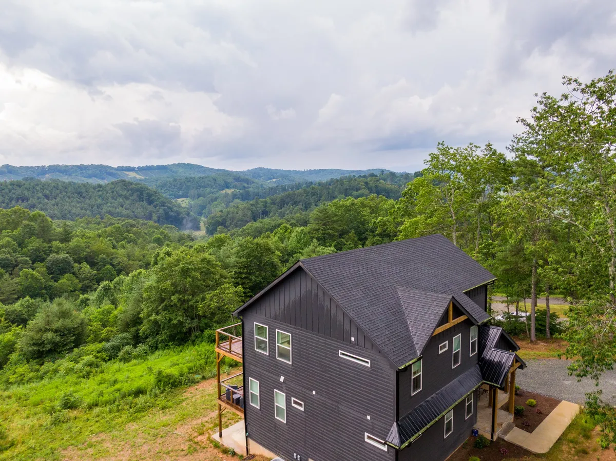The image showcases a modern, newly built house nestled among lush greenery, with expansive views of distant Blue Ridge mountains under a tranquil sky. The home’s inviting porch faces stunning sunset vistas, offering a serene spot to relax and immerse in nature.